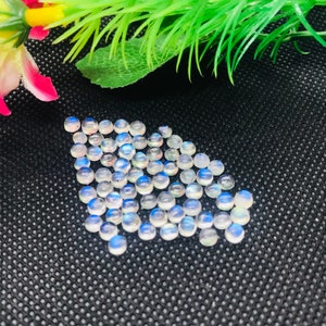 May include: A collection of round, iridescent moonstone cabochons in various shades of white and blue. The stones are arranged in a loose pile on a black textured surface.