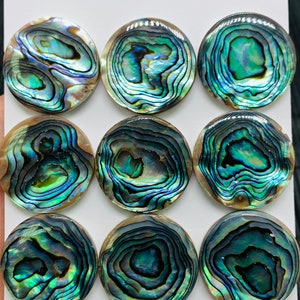 May include: Nine round iridescent abalone shell cabochons, each with a unique swirling pattern of green, blue, and brown. The cabochons are 25mm in diameter and are arranged in three rows on a white background. The text "25mm" and "9pcs" is written on the background.
