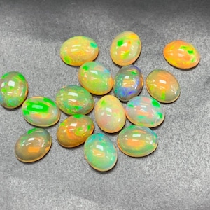 Puede incluir: Una colección de piedras preciosas de ópalo de forma ovalada en varios tonos de verde, amarillo y naranja. Los ópalos son iridiscentes y tienen una apariencia translúcida.