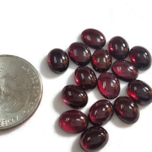 May include: A collection of 18 oval-shaped, red gemstones. The gemstones are smooth and polished, with a slight shine. They are arranged in a loose pile on a white surface.