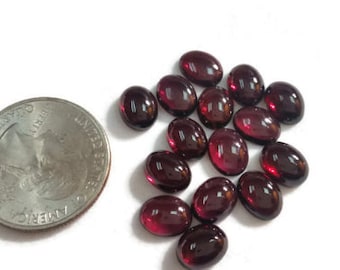 May include: A collection of 18 oval-shaped, red gemstones. The gemstones are smooth and polished, with a slight shine. They are arranged in a loose pile on a white surface.