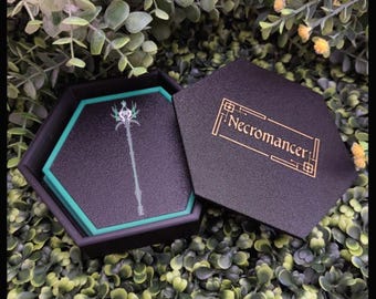 May include: A black hexagonal box with a teal border, containing a black coaster with a teal border and a green and white staff design. The box lid is black with gold lettering that reads "Necromancer".