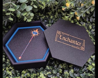 May include: A black hexagonal box with a blue trim and a black interior. The box is open and shows a black tray with a blue trim. The tray has a colorful illustration of a staff with a blue orb on top. The box lid is black with gold lettering that reads "Enchanter".