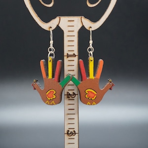 May include: Thanksgiving-themed earrings featuring hand-shaped turkeys with colorful fingers. The earrings are brown with red, orange, yellow, and green accents. The earrings are suspended from silver hooks and displayed on a wooden stand.