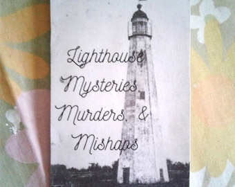 May include: A black and white postcard featuring a tall lighthouse. The text on the card reads "Lighthouse Mysteries, Murders, & Mishaps" in elegant cursive script. The lighthouse is set against a dark landscape.