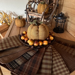 May include: A set of eight brown and tan plaid fabric placemats arranged in a circle around a plate of small brown and orange fruit. A large brown fabric pumpkin sits on top of the plate. Three smaller fabric pumpkins are visible in the background.