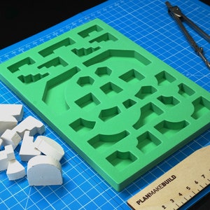May include: A green foam mold with various shapes for creating miniature buildings. The mold is on a blue cutting mat with a wooden ruler labeled "PlanMakeBuild" and a pair of silver compasses. There are several white foam pieces scattered on the mat.
