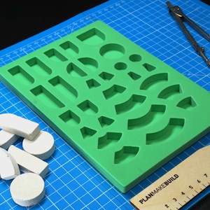 Puede incluir: Un molde de silicona verde con varias formas, incluyendo círculos, cuadrados, rectángulos y arcos. El molde está sobre una estera de corte azul con una regla de madera con la etiqueta "PLANMAKEBUILD". Hay formas de espuma blanca al lado del molde.