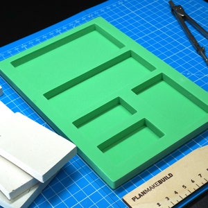 May include: A green foam tray with multiple rectangular cutouts, a white foam board, a wooden ruler with the text "PLANMAKEBUILD", and a silver compass on a blue cutting mat.