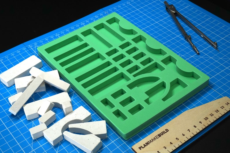 May include: A green silicone mold with various shapes and sizes, including rectangles, squares, and arches. The mold is on a blue cutting mat with a wooden ruler and a compass.