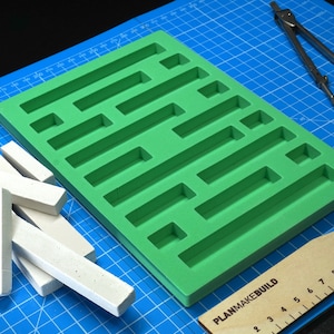 Könnte beinhalten: Grüne Silikonform mit rechteckigen Vertiefungen auf einer blauen Schneidematte. Ein Satz weißer rechteckiger Stäbe liegt verstreut auf der Matte. Ein Holzlineal mit dem Text "PlanMakeBuild" befindet sich in der unteren rechten Ecke. Ein Zirkel befindet sich in der oberen rechten Ecke.