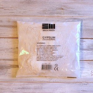 May include: A transparent bag containing white gypsum powder, marked "GYPSUM (CALCIUM SULPHATE)" by Brick Maker. The packaging features a warning and the product's art number, BMC-100.
