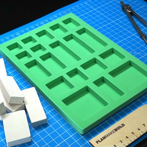 Könnte beinhalten: Eine grüne Schaumform mit 16 rechteckigen Fächern in verschiedenen Größen. Die Form liegt auf einer blauen Schneidematte mit einem Lineal und einem Zirkel daneben. Auf der Matte liegen auch mehrere weiße Schaumstoffblöcke.