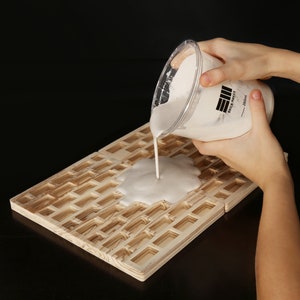 May include: A white, opaque liquid is being poured from a clear plastic container onto a wooden surface with a grid pattern. The container has a black label with the text "EII Brick Paste - 250ml".