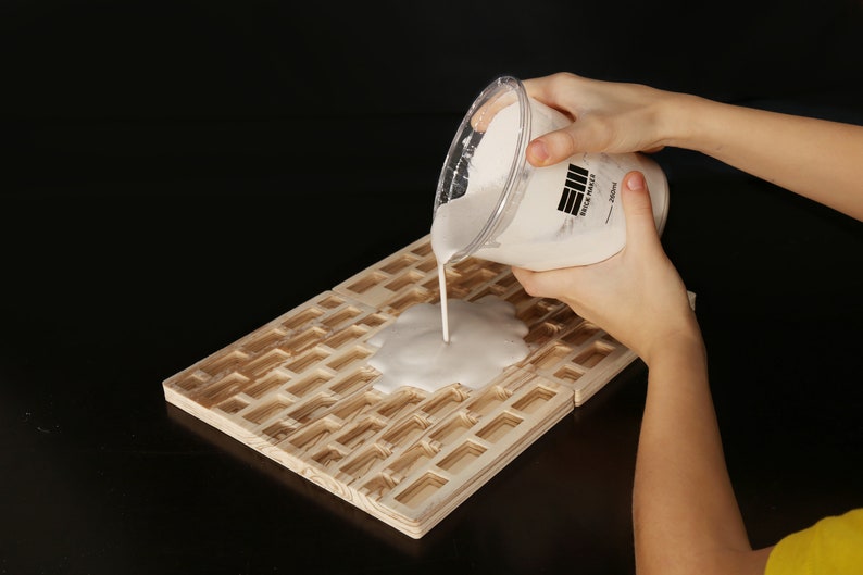 May include: A white, opaque liquid is being poured from a clear plastic container onto a wooden surface with a grid pattern. The container has a black label with the text "EII Brick Paste - 250ml".