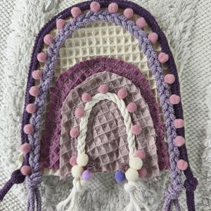 Könnte beinhalten: Ein handgefertigter Regenbogen-Wandbehang in Lila-, Lavendel- und Weißtönen. Der Regenbogen hat ein strukturiertes Waffelwebmuster und ist mit kleinen rosa Pompons verziert. Die Enden des Regenbogens haben geknotete Seildetails.
