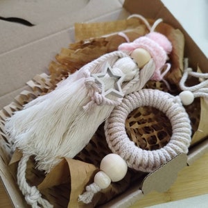 Könnte beinhalten: Eine Geschenkbox gefüllt mit handgefertigten Artikeln, darunter eine beige und weiße Quastenpuppe mit rosa Hut, ein beigefarbener Makrameering und Holzperlen.