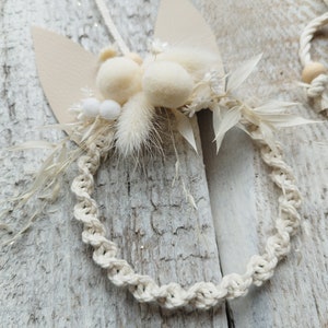 May include: A white macrame wreath with dried white flowers and fluffy white pom poms. The wreath is hanging from a white string on a light wood background.