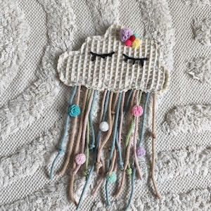 Könnte beinhalten: Cremefarbene, wolkenförmige Wanddekoration mit Waffelstruktur. Die Wolke hat geschlossene Augen und bunte Garnstränge mit Pompons. Die Garnstränge sind in den Farbtönen Rosa, Blau, Grün und Creme gehalten.