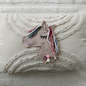 Könnte beinhalten: Ein beigefarbenes Kissen mit einer strukturierten Oberfläche und einer rosa-weißen Einhornkopf-Applikation mit buntem Garnhaar.