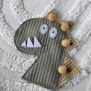 Könnte beinhalten: Handgefertigte Dinosaurier-Applikation aus olivgrünem Waffelstoff. Mit Filzzähnen, gestickten Augen und beigen Filzakzenten. Der Rücken des Dinosauriers ist mit kleinen, beigen Filzkugeln verziert. Ein charmantes Deko-Element.