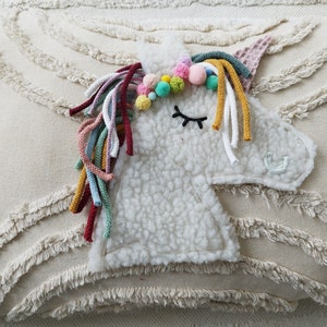 May include: A white plush unicorn pillow with a colorful yarn mane and a closed eye. The pillow is on a cream-colored background with a textured fringe.