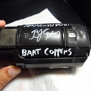 Op de afbeelding: Een zwarte Sony camcorder met een zilverkleurig microfoongedeelte van gaas en een zilverkleurige schuifschakelaar. Het apparaat is voorzien van witte handgeschreven tekst "BART COPPENS" en een handtekening, samen met de datum "10/26/22-14/06".