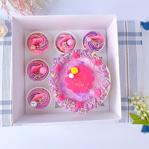 Könnte beinhalten: Eine weiße Box mit einem dekorierten Kuchen und Cupcakes. Der Kuchen ist rund mit lila Zuckerguss und einem rosa "Happy Birthday"-Schild. Die Cupcakes haben rosa und lila Zuckerguss mit dekorativen Elementen. Die Box befindet sich auf einem blau-weiß gestreiften Tuch.