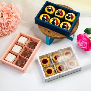 May include: Three rectangular gift boxes containing assorted pastries. One dark blue box holds six yellow fruit-topped cakes. A light pink box contains three white square treats and three round chocolate items. A white marble-patterned box holds six jam-filled cookies.