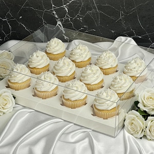 Op de afbeelding: Een dozijn cupcakes in een doorzichtige doos, elk met een werveling van witte frosting en zilveren sprinkles. De cupcakes staan op een witte tray, omringd door witte rozen op een witte satijnen ondergrond.