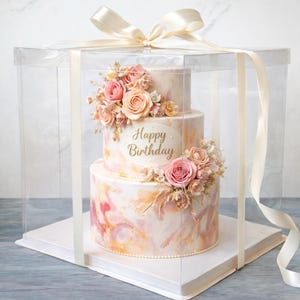 May include: A three-tiered birthday cake with pink and peach floral decorations, presented in a clear box tied with a cream ribbon. The cake has a marble-like design and the words "Happy Birthday" are displayed on the front.