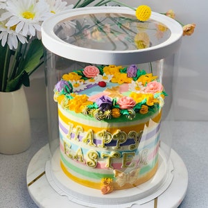 Peut inclure: Un gâteau coloré décoré de fleurs et des mots "Happy Easter" dans un récipient en plastique transparent sur un support à gâteau blanc.