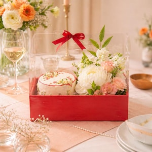 Könnte beinhalten: Eine rote Box mit einem transparenten Deckel enthält einen kleinen Kuchen und ein Blumenarrangement. Der Kuchen ist weiß mit floralen Dekorationen. Die Blumen umfassen weiße Pfingstrosen, rosa Rosen und Grünzeug. Oben auf der Box ist ein rotes Band gebunden.