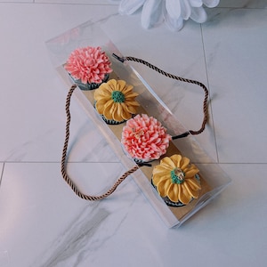 Op de afbeelding: Een doorzichtige doos met vier cupcakes met bloemvormige glazuur. De cupcakes hebben roze en gele glazuur met groene en gouden accenten. De doos heeft een bruin touwhandvat.