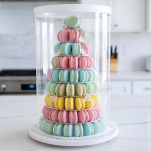 May include: A colorful tower of macarons in a clear display case. The macarons are arranged in a pyramid shape, with layers of pink, blue, yellow, and green. The display case sits on a white, round base.