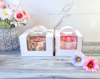 May include: Two white cardboard cake boxes with clear plastic windows, each containing a small decorated cake. The left cake features a multi-colored floral pattern, while the right cake is topped with pink frosting swirls and small sprinkles.