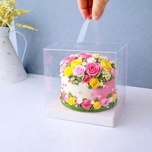 May include: A round cake with pink frosting and a floral design of yellow, pink, and white roses. The cake is in a clear plastic box with a handle.