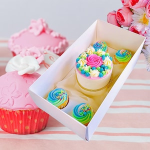 Könnte beinhalten: Eine weiße Box gefüllt mit Cupcakes. Die Cupcakes sind mit buntem Frosting und Streuseln verziert. Die Box steht auf einer rosa-weißen gestreiften Oberfläche.