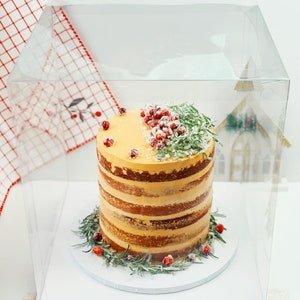 May include: A tall, multi-layered naked cake with light orange frosting between the layers. The top is decorated with clusters of red cranberries and green rosemary sprigs dusted with white sugar, all enclosed in a clear plastic box.