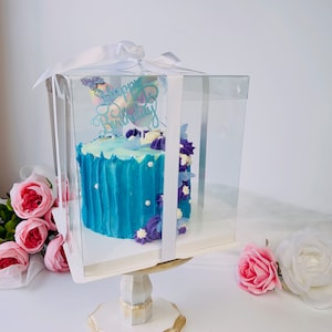 May include: A blue frosted cake with white pearl accents and purple floral decorations, presented in a clear box tied with a white ribbon. The cake has a 'Happy Birthday' topper. The cake sits on a decorative stand.