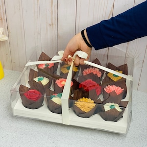 May include: A clear plastic bakery box filled with a dozen cupcakes. Each cupcake is in a brown paper cup and topped with colorful buttercream flowers. The box is tied with a cream-colored ribbon.
