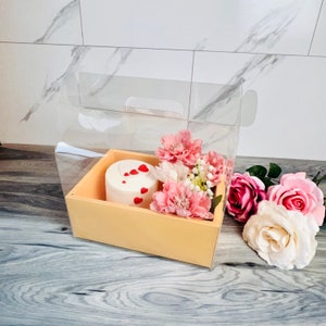 Könnte beinhalten: Ein kleiner, runder weißer Kuchen, verziert mit roten Herzen, befindet sich in einer hellbraunen Holzkiste, umgeben von rosa und weißen Blumen. Die Kiste befindet sich in einem transparenten Kunststoffbehälter mit einem Griff. Mehrere rosa und weiße Rosen liegen neben der Kiste.