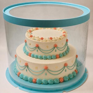 May include: A two-tier white frosted cake inside a clear cylindrical display box with a light blue base and lid. The cake features light blue piped swags, coral-colored rosettes, and small gold edible beads decorating the edges and sides.