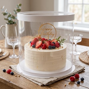 May include: A birthday cake with white frosting, decorated with fresh berries and a gold "Happy Birthday" topper. The cake is enclosed in a clear plastic container with a white lid and base, presented on a wooden table.