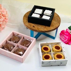 Op de afbeelding: Een assortiment lekkernijen in decoratieve dozen. De afbeelding toont vier dozen: een zwarte doos met witte vierkanten, een roze doos met chocoladelekkernijen en een doos met marmerpatroon met koekjes. Een kleine houten tafel en roze bloemen staan ook op de foto.