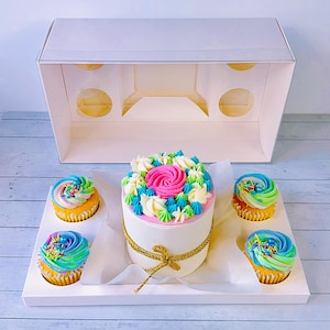 Könnte beinhalten: Eine weiße Kuchenbox mit einem runden Kuchen, der mit rosa, blauem und weißem Frosting verziert ist. Vier Cupcakes sind um den Kuchen herum angeordnet, jeder mit blauem, grünem und gelbem Frosting. Die Cupcakes sind mit Streuseln verziert.