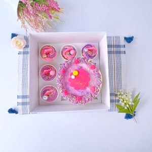 Könnte beinhalten: Eine weiße Box enthält einen kleinen Kuchen und sechs Cupcakes, alle mit rosa und lila Zuckerguss verziert. Der Kuchen hat ein rosa Etikett mit der Aufschrift "Happy Birthday". Die Cupcakes haben verschiedene Beläge, darunter rosa Federn und Perlen.