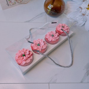 May include: A clear plastic box with a handle contains four pink cupcakes with green sprinkles. The cupcakes are arranged in a row and are decorated with a flower design.