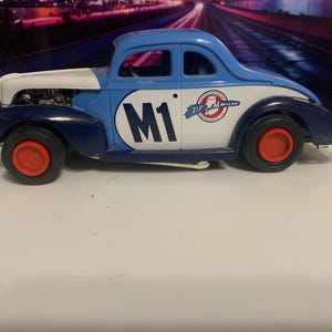 May include: A vintage-style toy car in blue, white, and navy with red wheels. The car features the number "M1" on the side and "Zip Plus Mileage" branding. The car is a classic coupe design.