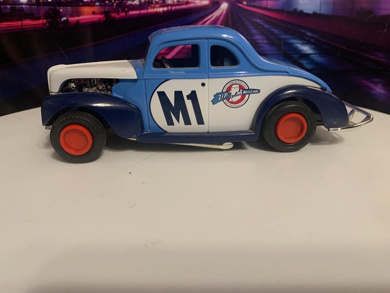 May include: A vintage-style toy car in blue, white, and navy with red wheels. The car features the number "M1" on the side and "Zip Plus Mileage" branding. The car is a classic coupe design.
